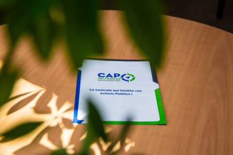 Sur une table est posé le catalogue de CAP Territoires. Comme inscription on peux lire : La centrale qui facilite os Achats Publics !