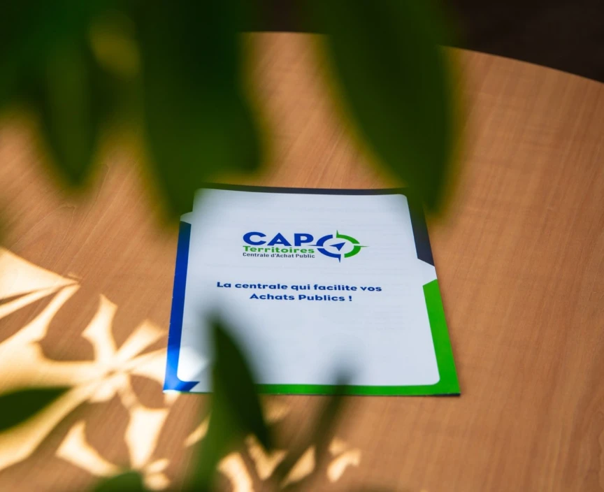 Sur une table est posé le catalogue de CAP Territoires. Comme inscription on peux lire : La centrale qui facilite os Achats Publics !